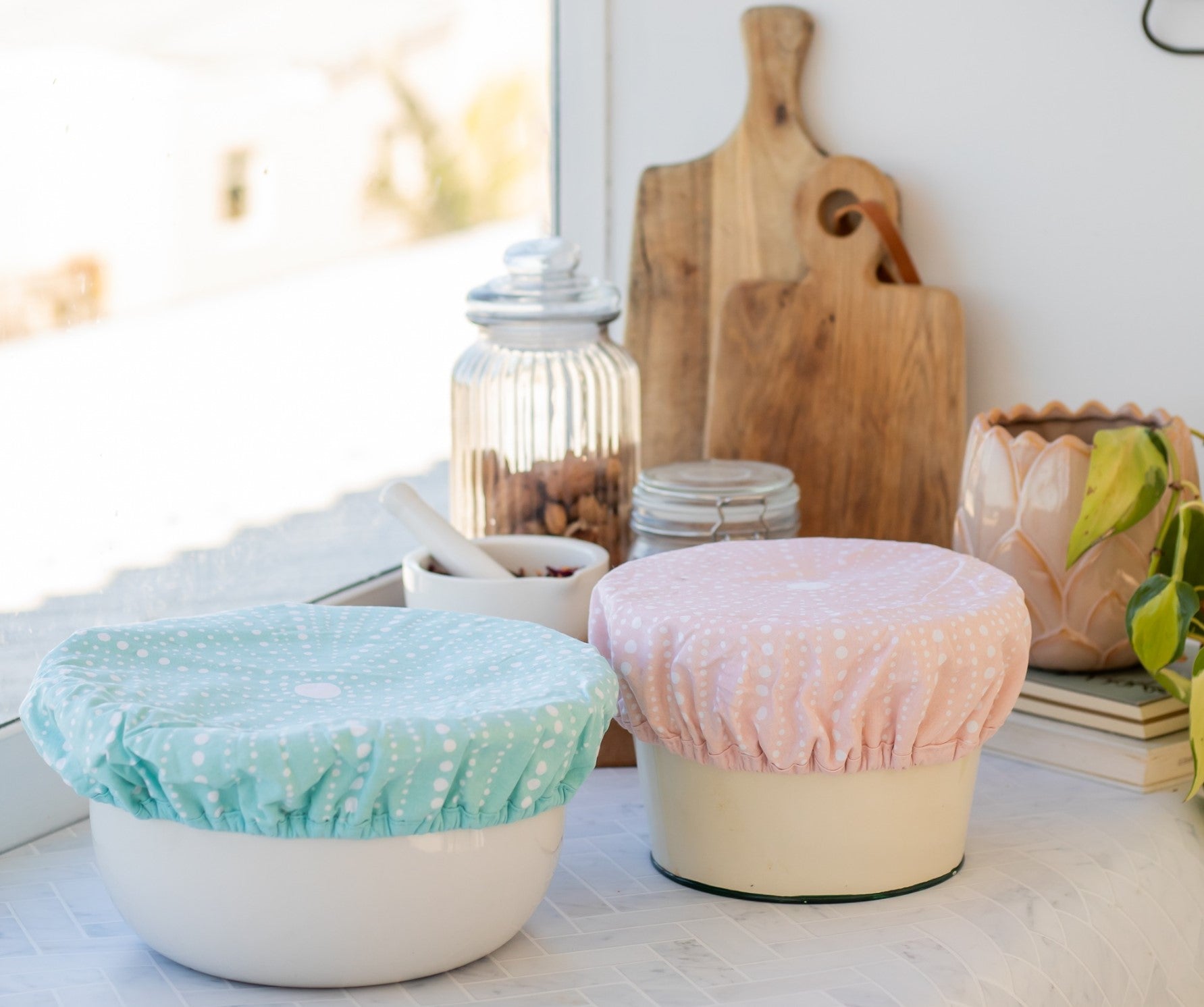 Two bowls on a kitchen counter are topped with Kina Bowl Covers (Set of 2), featuring pastel, food-safe certified, reusable cotton outers. Behind them: wooden cutting boards, a glass jar, a plant, and a ceramic container.