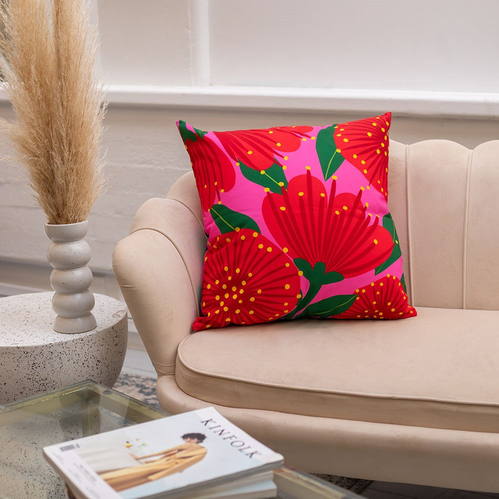 The Bright Botanical Pink Pohutukawa Cushion Cover, featuring a red floral polyester print on pink, sits on a light beige sofa. A pampas grass vase and magazine enhance the modern, minimal setting for indoor and outdoor use.