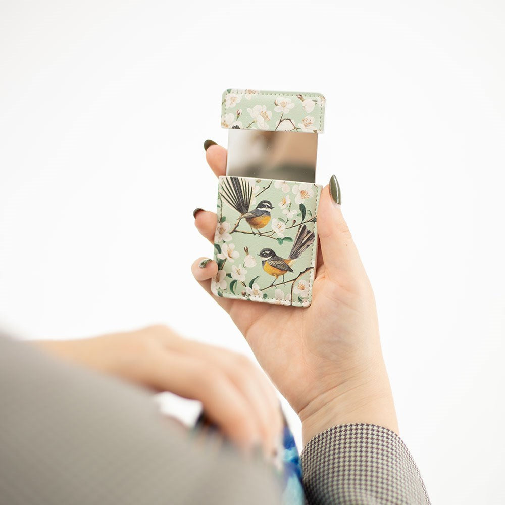 A person with dark nail polish holds an open Chatty Fantails Pocket Mirror, featuring a mint green vegan leather case with bird and floral designs. The inside of the case is visible against a plain white background.