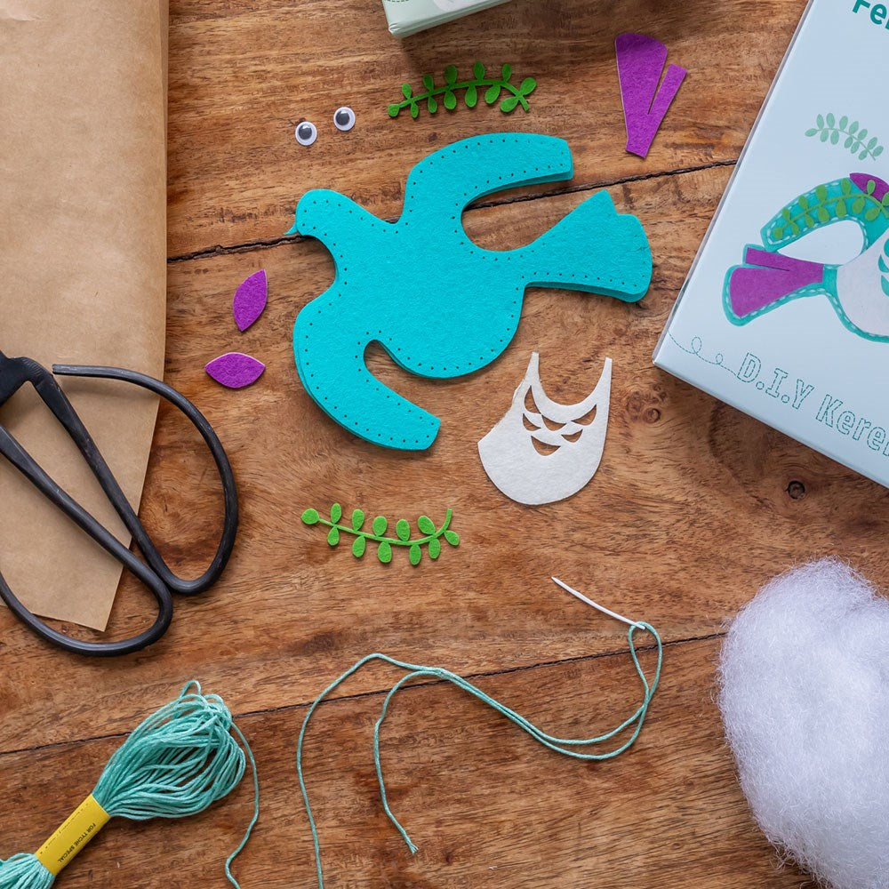 Kereru Felt DIY Kit materials on a wooden table: pre-cut felt pieces, needle with thread, stuffing, scissors, googly eyes, and embroidery floss—the perfect start for creative projects and heartfelt gifts.