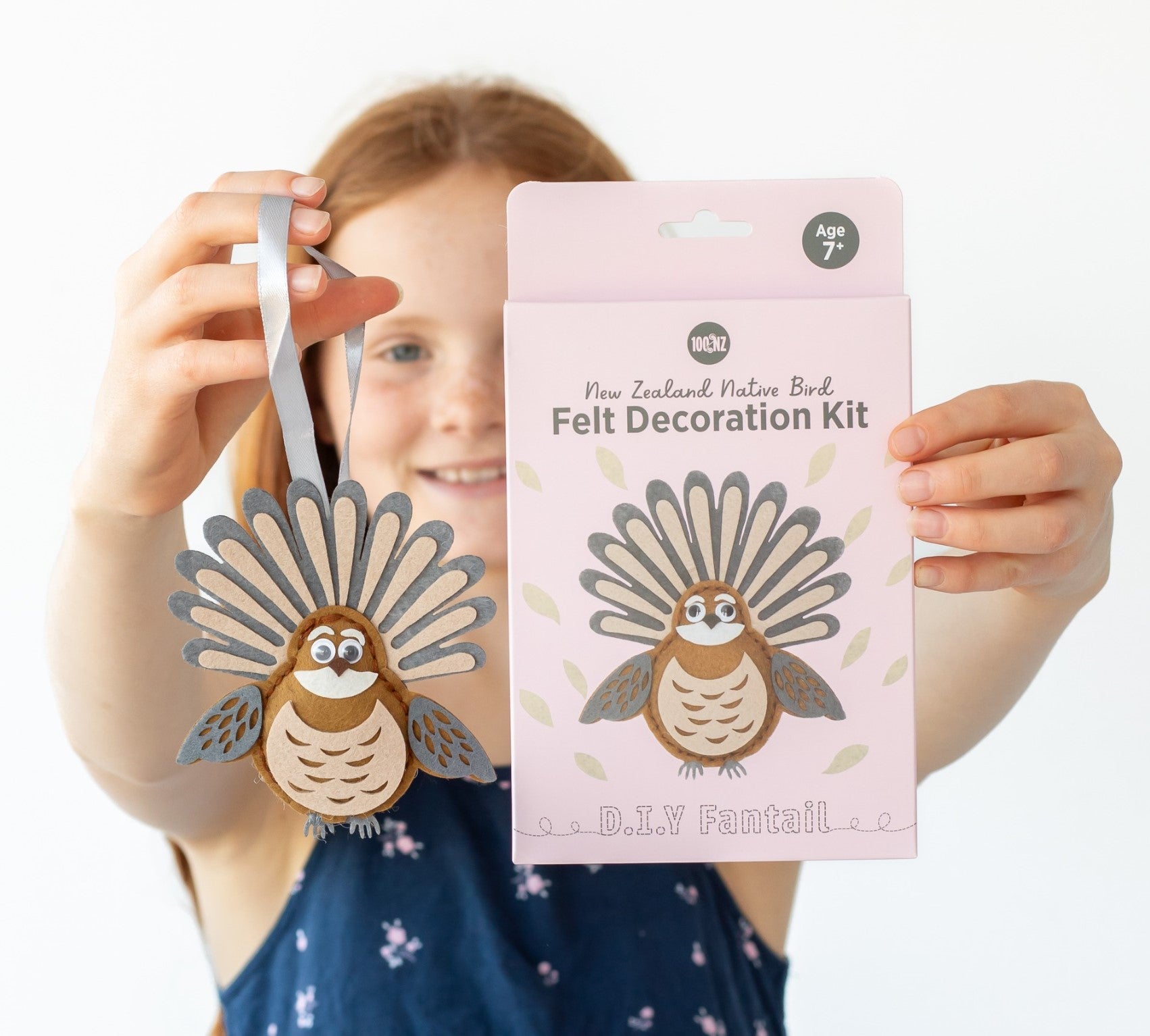 A child holds up a felt bird ornament in one hand and the Fantail Felt DIY Kit box in the other, proudly displaying their handmade creation that perfectly matches the kit’s design.