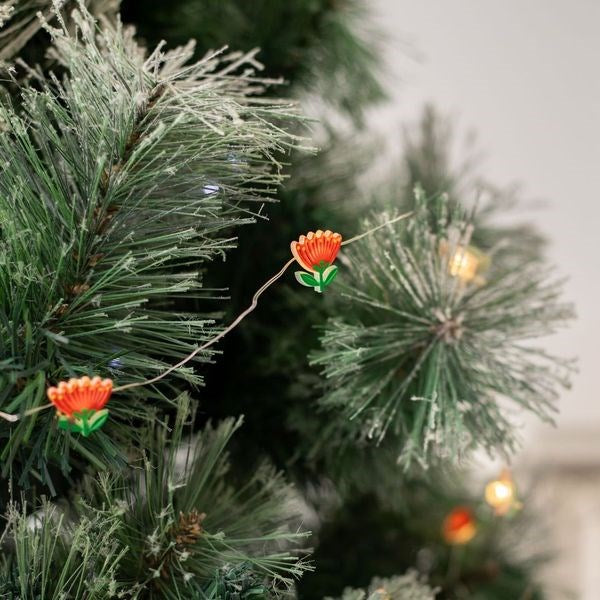 Close-up of a green Christmas tree with Pohutukawa LED Lights—battery-powered, warm yellow bulbs and red flower shapes. Designed in New Zealand, these lights add festive charm among frosted needles.