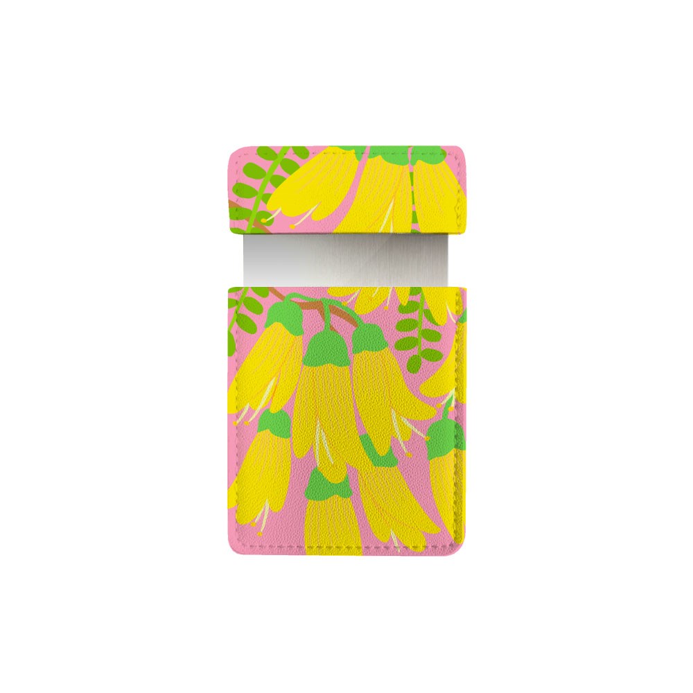 The Bright Botanical Kowhai Pocket Mirror features yellow and green flowers with pink accents on a pink background. Crafted from vegan leather, it has an open top flap and a light gray interior.