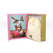 The Angie Dennis - Deeds not Words Jigsaw Puzzle comes in a pink box featuring whimsical stacked teacups, birds, and a rose inside the cover. It celebrates the suffragette movement and includes a cream drawstring bag with black text.