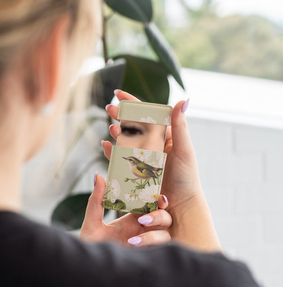 A person holds the Birds Botanics Rifleman Pocket Mirror, featuring a floral and bird design on the back—an ideal handbag accessory. A plant and white tiled wall can be seen in the background.