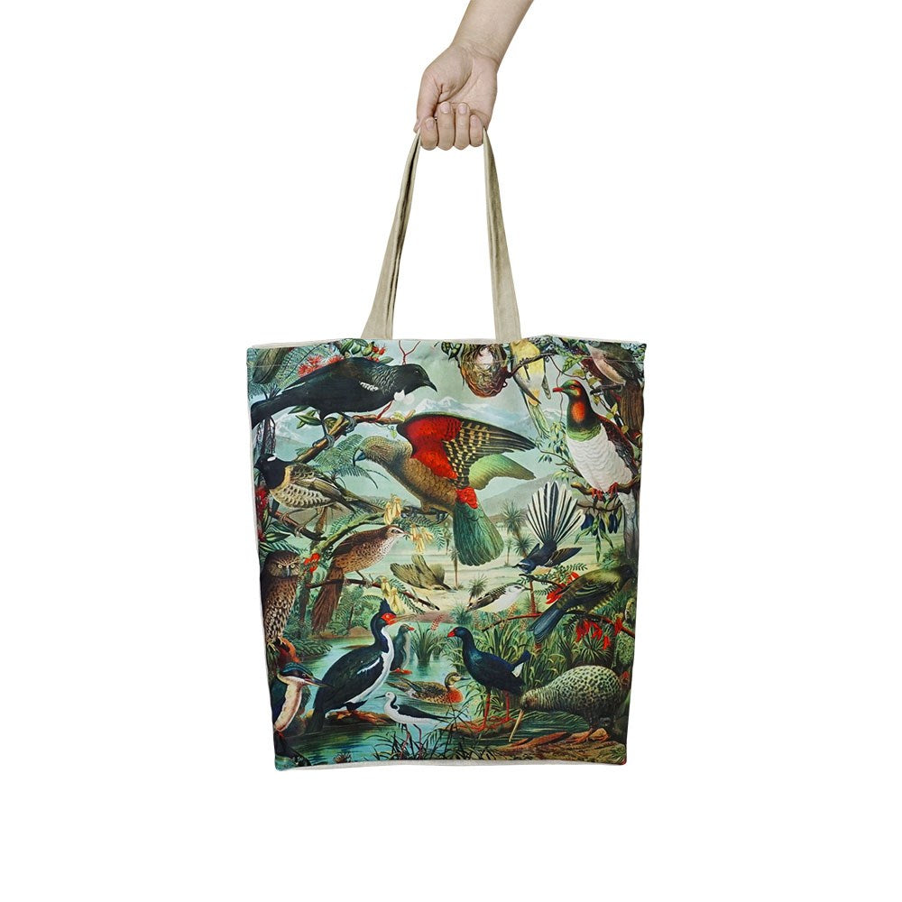 A hand holds the NZ Native Birds Shopping Bag, a lightweight tote with beige handles and a vibrant, detailed print of native birds in a natural setting.