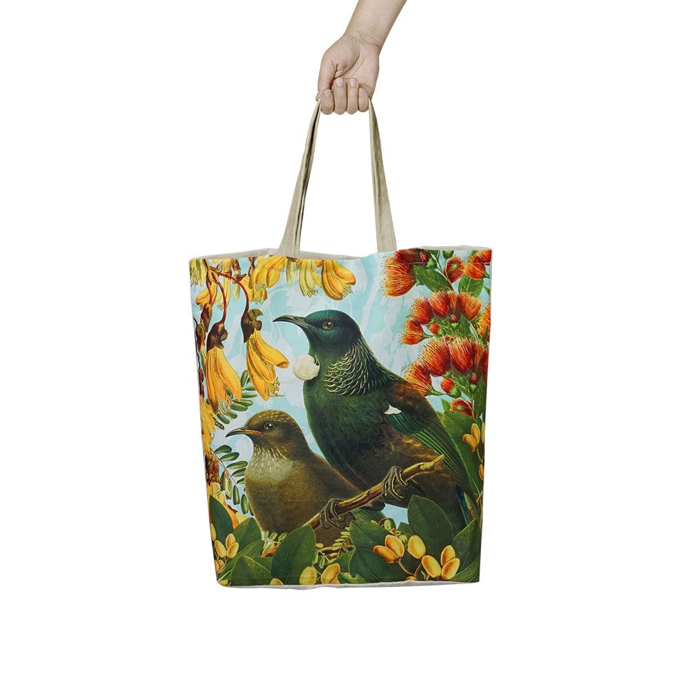 A hand holds the Botanical Tui Shopping Bag, a lightweight, reusable tote featuring two birds among yellow and red flowers with green leaves on a light blue background with cloud-like shapes.