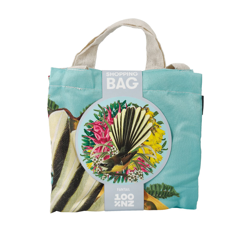 The Botanical Fantail Shopping Bag features beige handles, a light blue background, and a vibrant bird with flowers. Affordable and stylish, its front label reads SHOPPING BAG FANTAIL 100% NZ.
