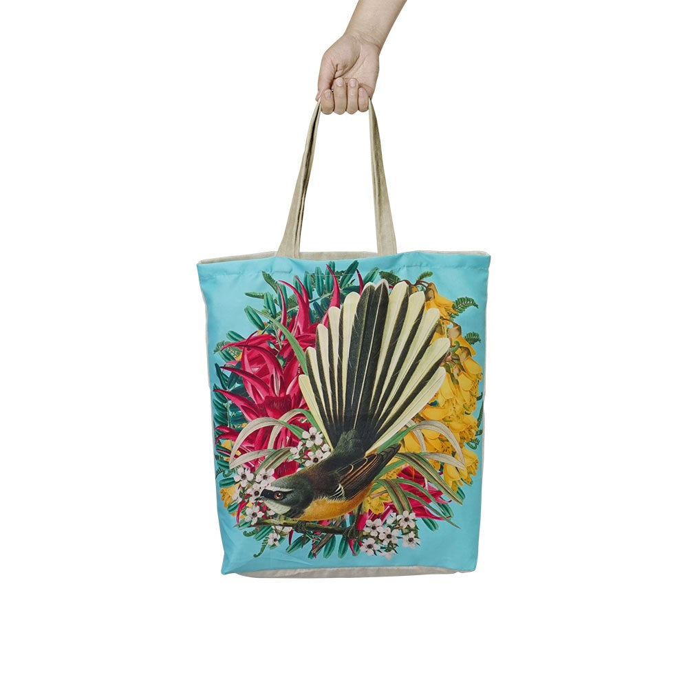A hand holds the affordable Botanical Fantail Shopping Bag, a reusable tote with beige handles featuring a bird with a fanned tail and colorful flowers on a light blue background.