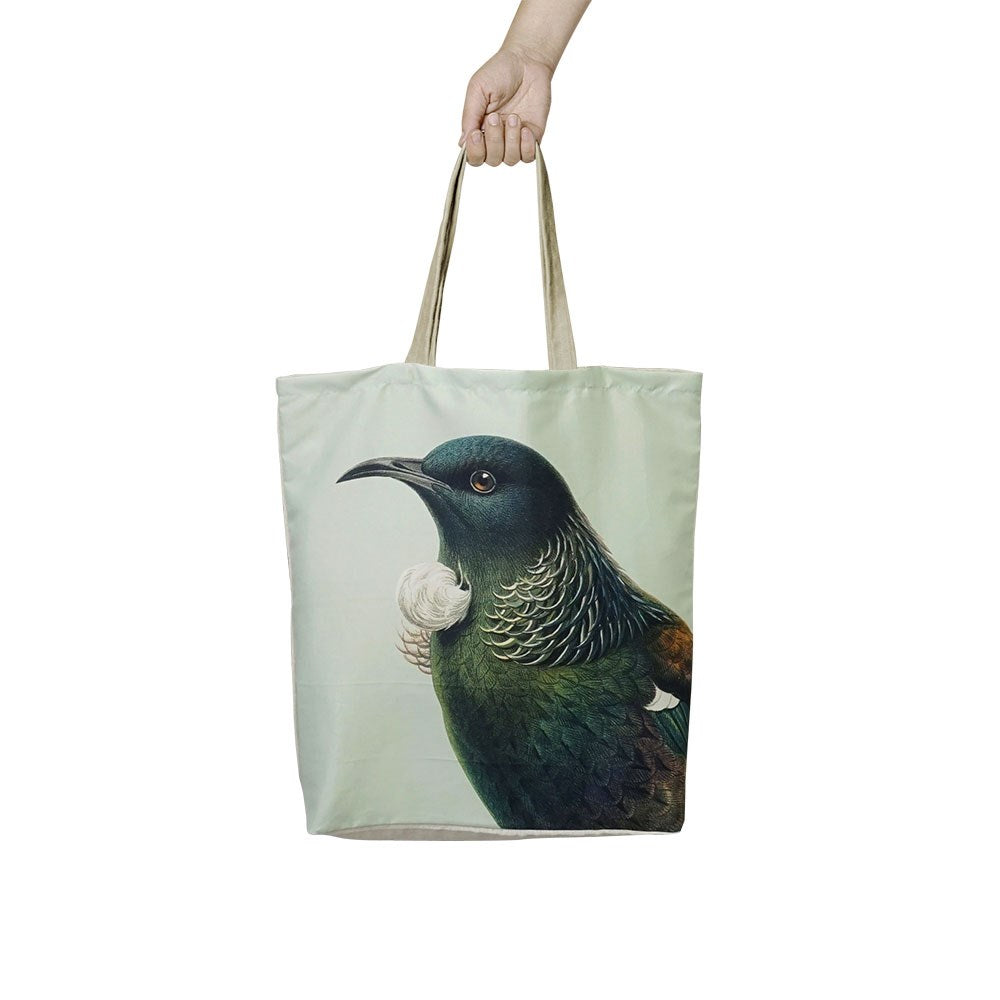 A hand holds the affordable, reusable Hushed Tui Shopping Bag, featuring a realistic tūī bird with iridescent green and blue feathers and a white neck tuft on a pale background.