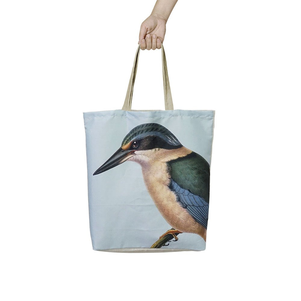 A hand holds the Hushed Kingfisher Shopping Bag, featuring beige handles and a kingfisher illustration on the front, with a plain light background.