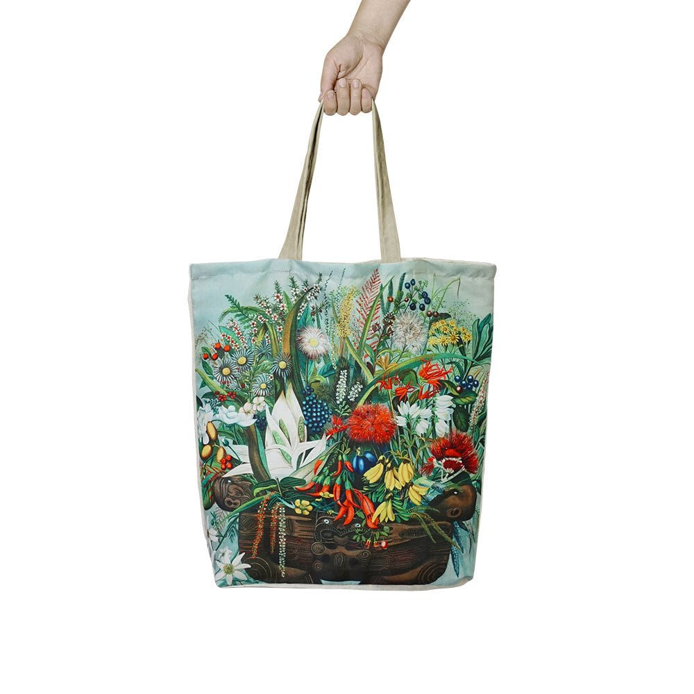 A hand holds the NZ Native Flowers Shopping Bag—an affordable tote with a beige handle and a colorful illustrated design of native flowers and plants on a light background, ideal for stylish, reusable shopping.