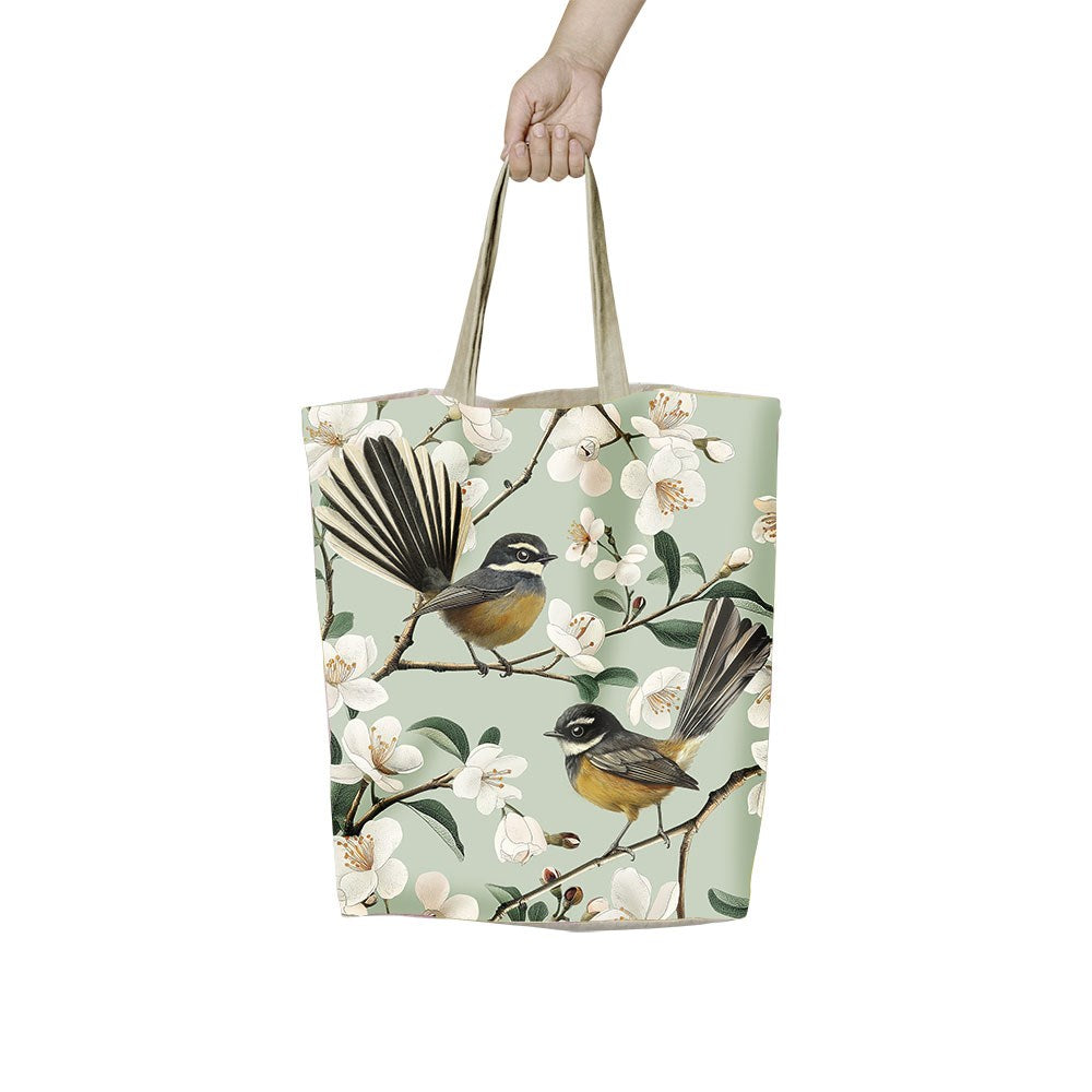 A hand holds the Chatty Fantails Shopping Bag—an affordable beige tote with two birds on blossoming branches against a light green background, offering a stylish and reusable option for everyday errands.
