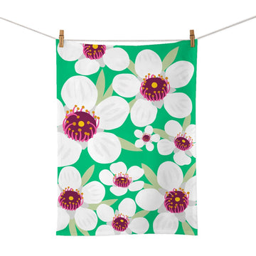 The Bright Botanical Manuka Tea Towel, made of cotton with a green background, features large white flowers with pink and yellow centers and green leaves, clipped to a clothesline.