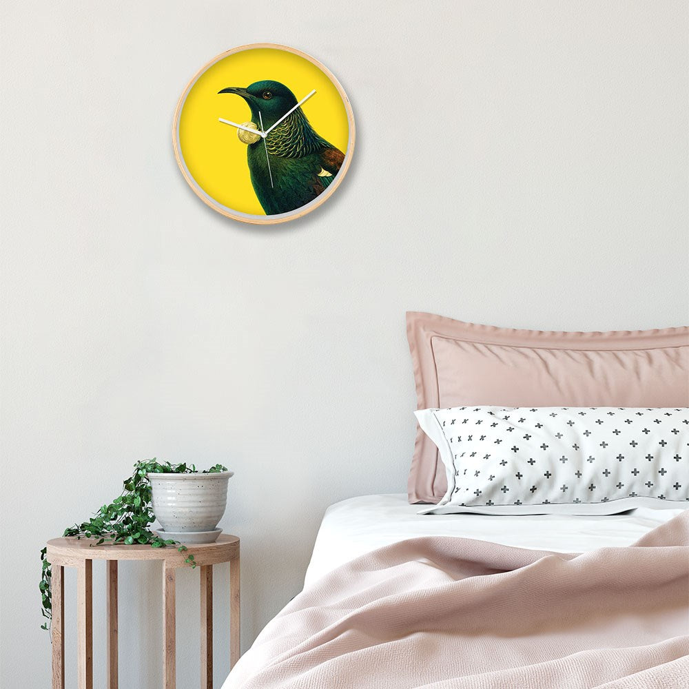 A bedroom with a neatly made bed, blush pink bedding, and a black cross pillow. A small plant sits on a wooden stool beside the bed. Above hangs the Bright Tui Wooden Frame Clock with silent movement and a bird illustration.