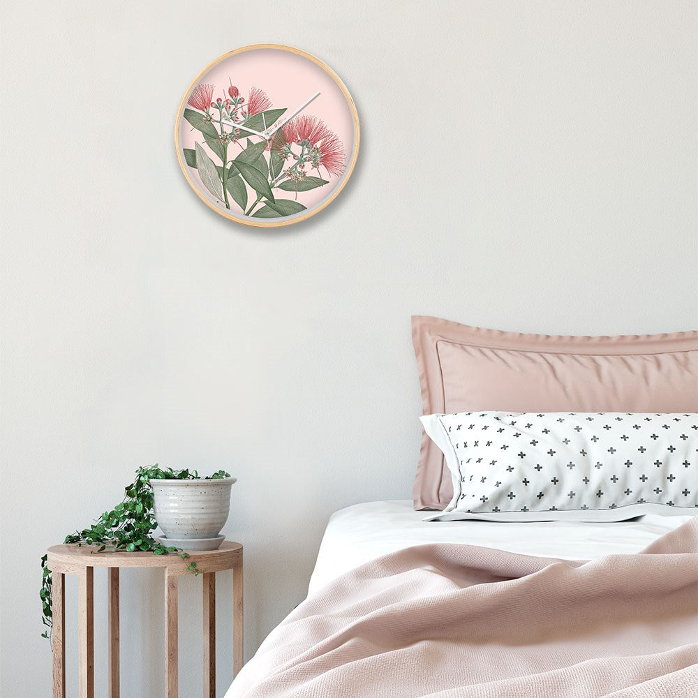 A neatly made bed with pink bedding sits by a small wooden side table topped with a trailing plant. Above the bed, a Vintage Botanical Pohutukawa Wooden Frame Clock hangs on the pale pink wall.