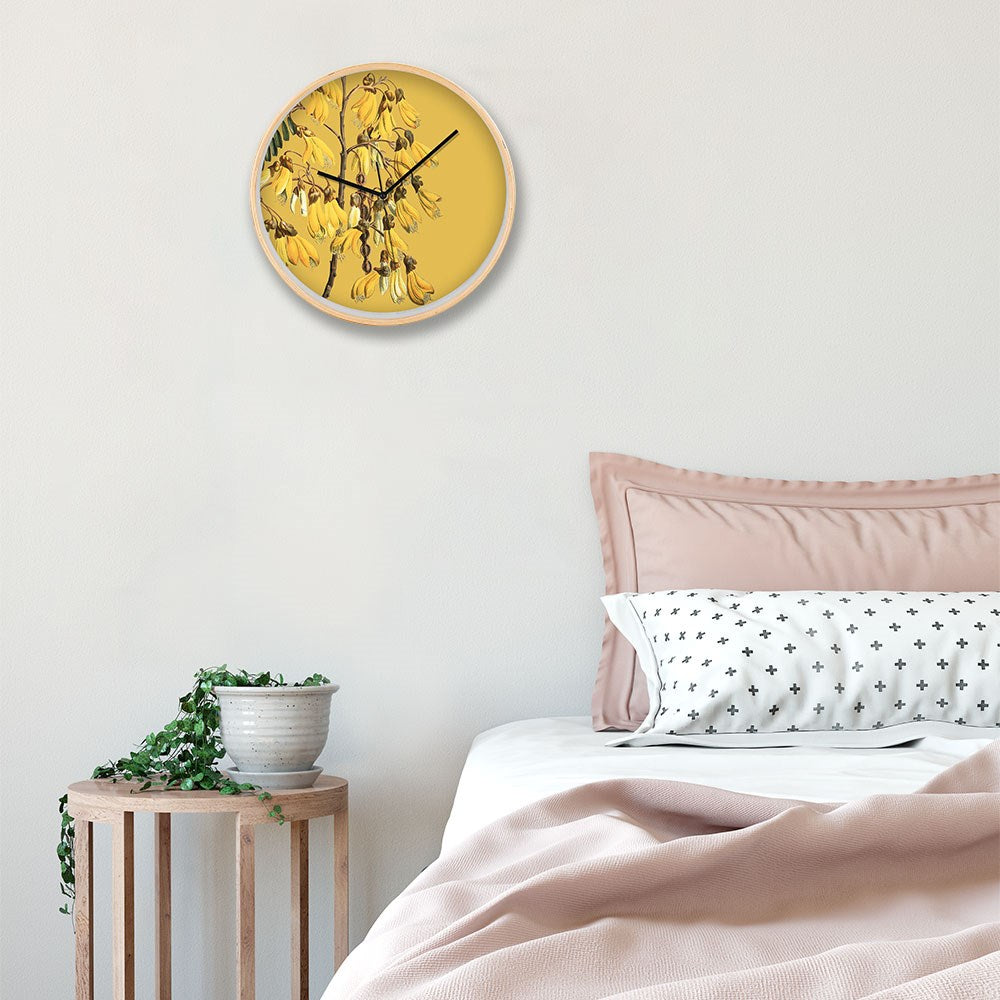 A wooden stool with a potted trailing plant sits by a bed with pink bedding and a white pillow with black crosses. Above, a Vintage Botanical Kowhai Wooden Frame Clock adds color to the light wall.