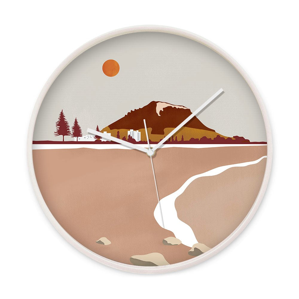 The Mt Maunganui Landmark Wooden Frame Clock features a minimalist New Zealand landscape with mountains, trees, and buildings under an orange sun. It has silent movement and white hands over a stream, rocks, and an earthy backdrop.