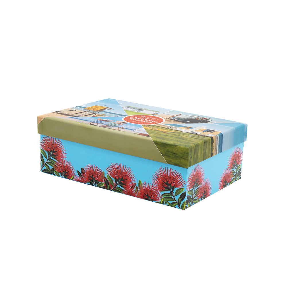 A rectangular cardboard box with a lid, adorned with images of red flowers and scenic landscapes inspired by Graham Young artworks, holds the Box of Six NZ Summer Christmas Baubles. The box is set against a plain white background.