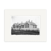 Pukekohe Primary 1910 - HI199: Black-and-white photo of a single-story building with large windows, gabled roofs, and chimneys, surrounded by a white fence and grass, mounted on archival quality matte paper.
