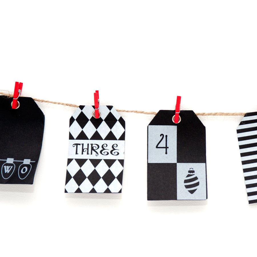 Four geometric black and white paper tags with numbers are clipped with red clothespins to string, creating a charming Advent Calendar with a craft style, displayed against a white background.