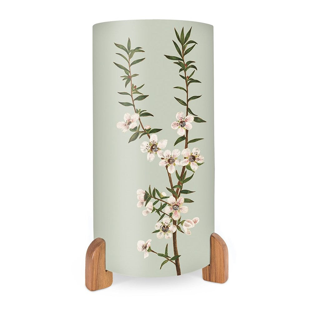 The Vintage Botanical Manuka Table Lamp is a cylindrical lamp with a pale green shade showcasing a vibrant New Zealand flowering branch, supported by two wooden feet.