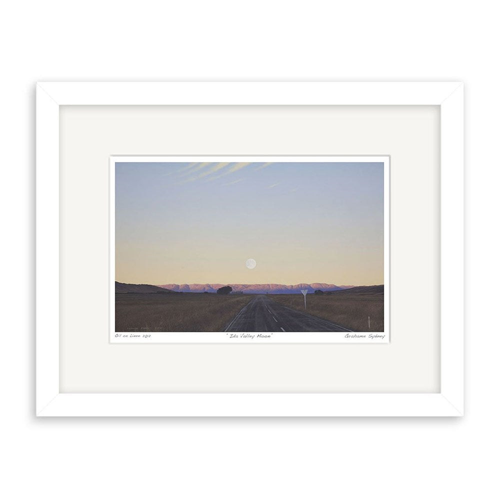 The Grahame Sydney - Ida Valley Moon Small Art Print: Second Frame features a paved road leading to distant mountains beneath a dusky sky with the moon on the horizon, double-matted and set in a sleek white frame.