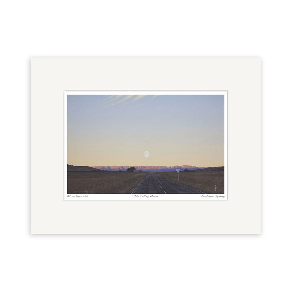 The Grahame Sydney - Ida Valley Moon Small Art Print features a framed landscape of a straight road toward distant mountains at sunset or sunrise, with a large low moon and soft pastel tones, inspired by Grahame Sydney prints.