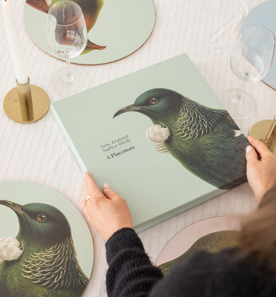 A person holds a box labeled Hushed Birds Box of 6 placemats with an illustration of a green native bird. On the table are wine glasses, matching bird-themed placemats, and a candle.