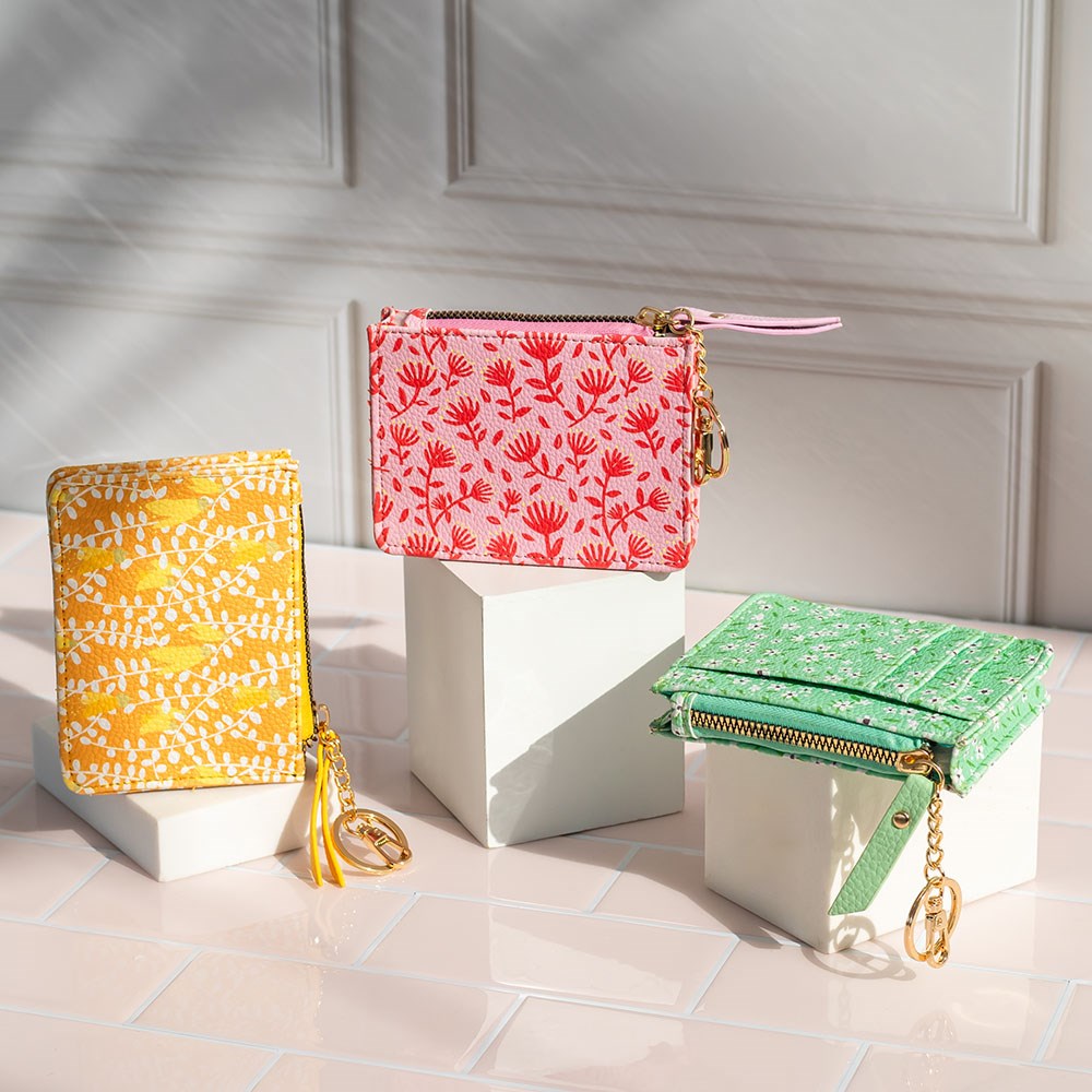 Three Manuka Pattern Coin Purses—yellow, pink, and green—crafted from vegan leather with gold keychain and zipper, are displayed on white blocks against a light tiled background.