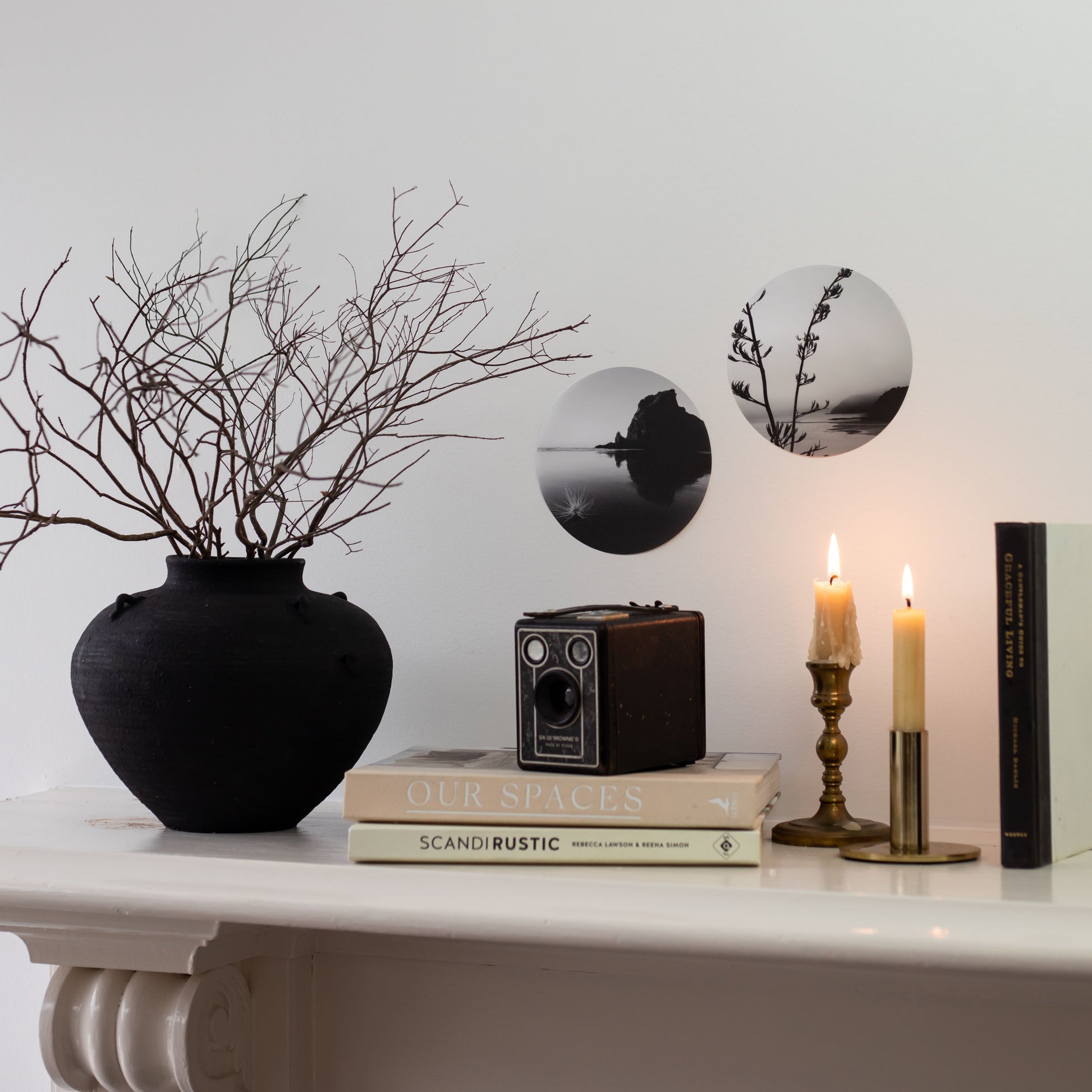 A black vase with bare branches, a vintage camera, stacked books, two lit brass candles, and the Piha Art Spot—matte fabric black-and-white circular wall art—style a white mantle against a white wall.