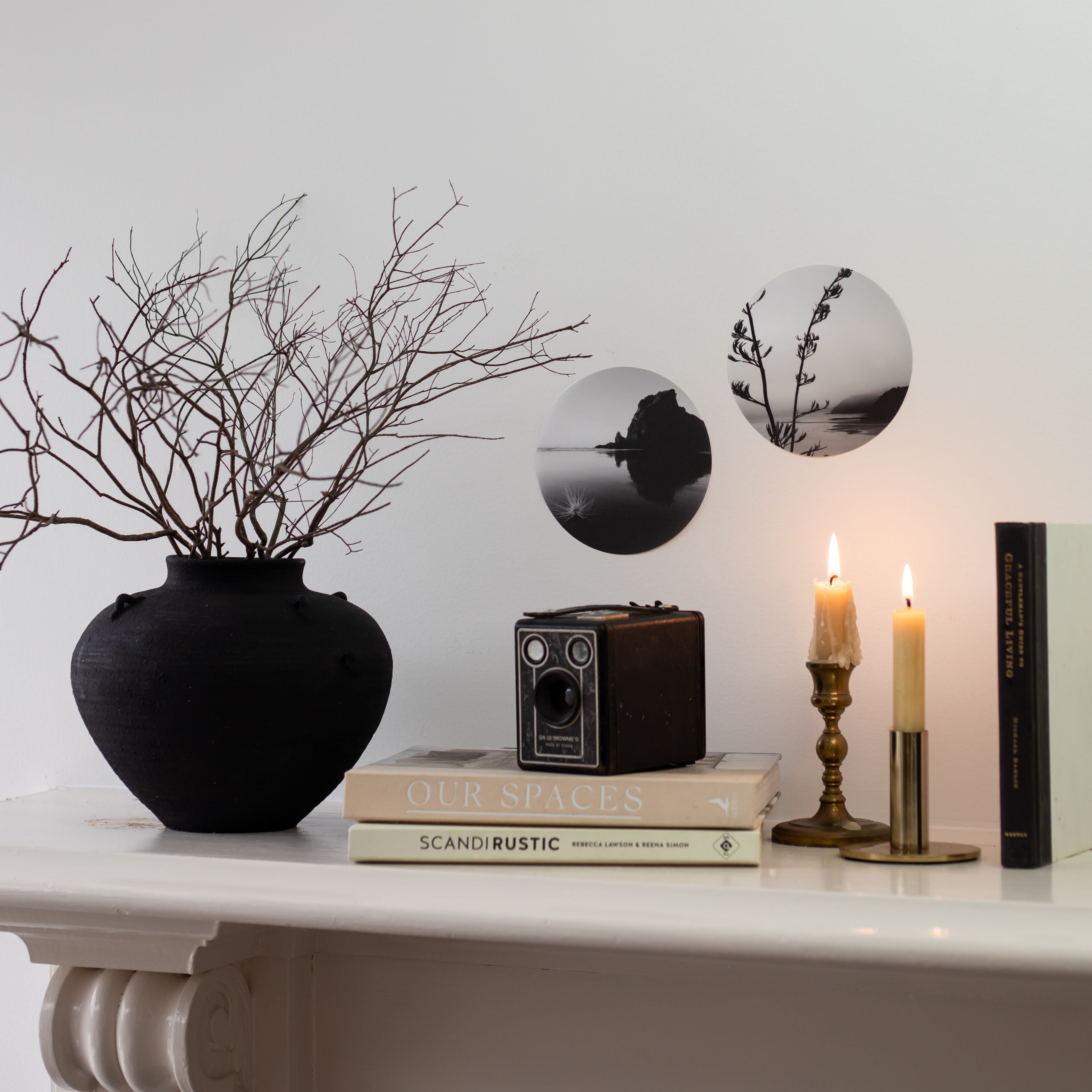 A black vase with bare branches, a vintage camera, stacked books, two lit brass candles, and the Piha Art Spot—matte fabric black-and-white circular wall art—style a white mantle against a white wall.