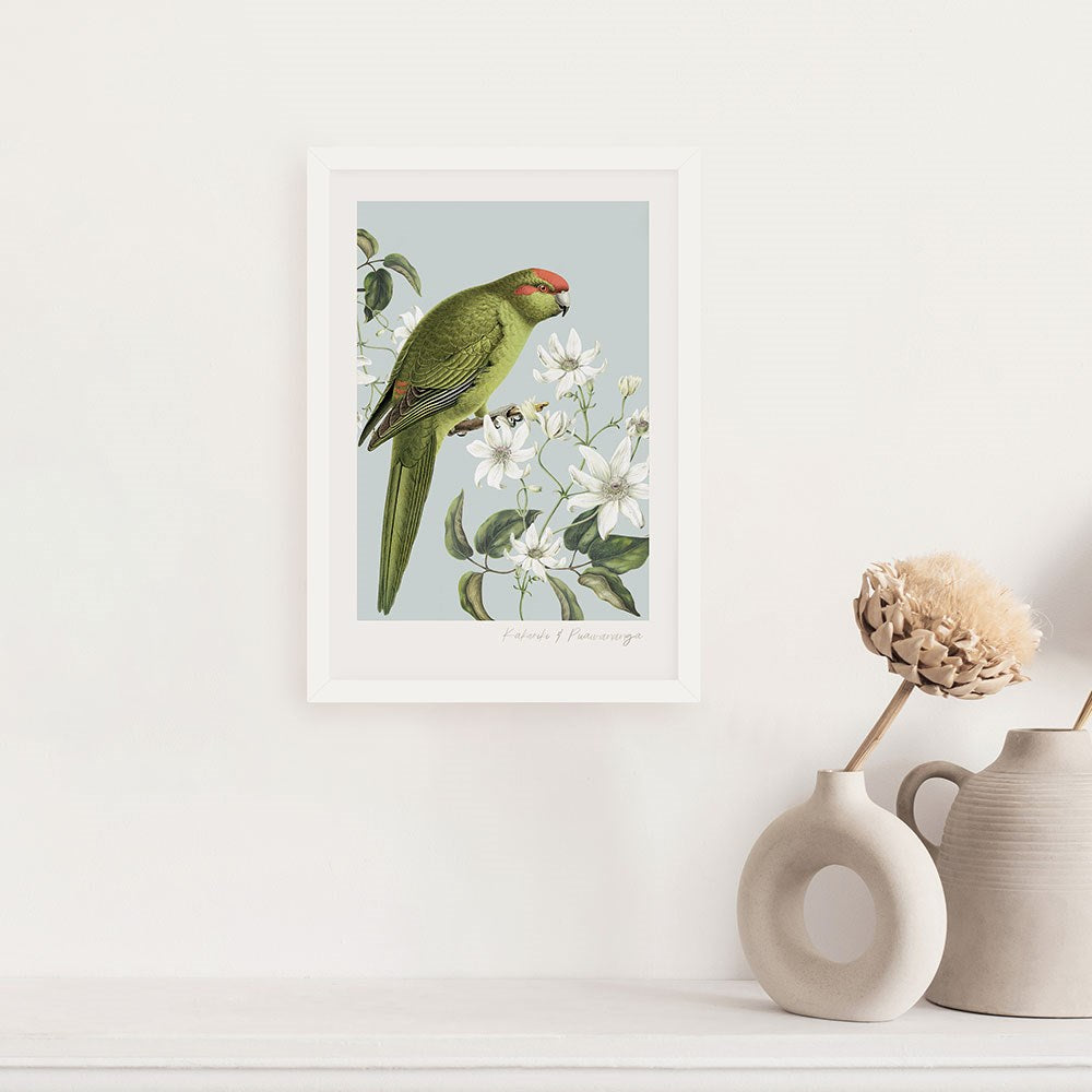 The Birds & Botanics Kakariki Print, a framed vintage-style illustration of a green parrot on white flowering branches, hangs above a light surface with two beige vases and dried flowers.