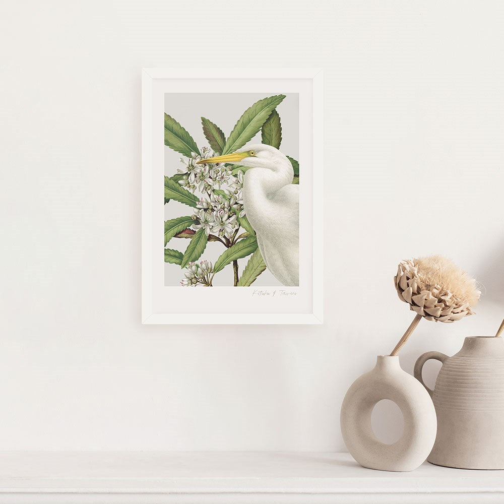 The Birds & Botanics Heron Print is a framed vintage-style botanical illustration featuring a white heron among green leaves and white flowers, perfect for hanging above a light surface with neutral vases and dried blooms.