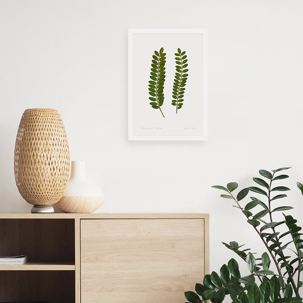 A light wood cabinet with two vases, one woven and one white, sits against a white wall. Above it hangs the framed Flora Kowhai Leaves A3 Print on art paper. A leafy green plant is in the lower right corner.