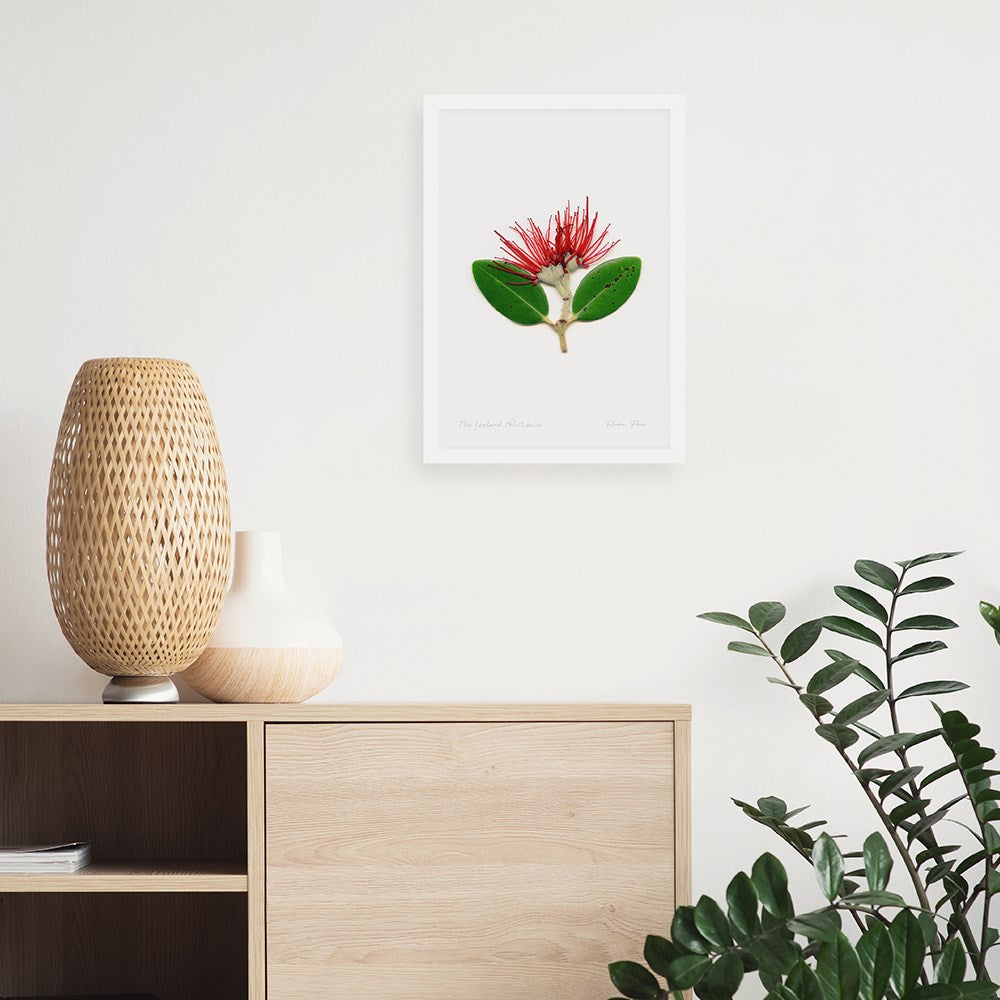 A light wood sideboard with two vases stands against a white wall. Above, the Flora Pohutukawa Portrait A3 Print displays a red and green flower. To the right of the sideboard sits a leafy green plant.
