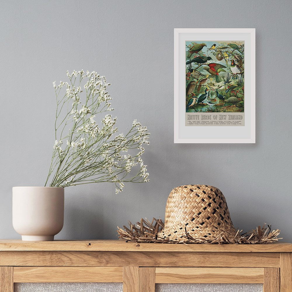 A straw hat and a white vase with babys breath sit on wood below an NZ Native Birds Print, framed and displayed on a light gray wall.