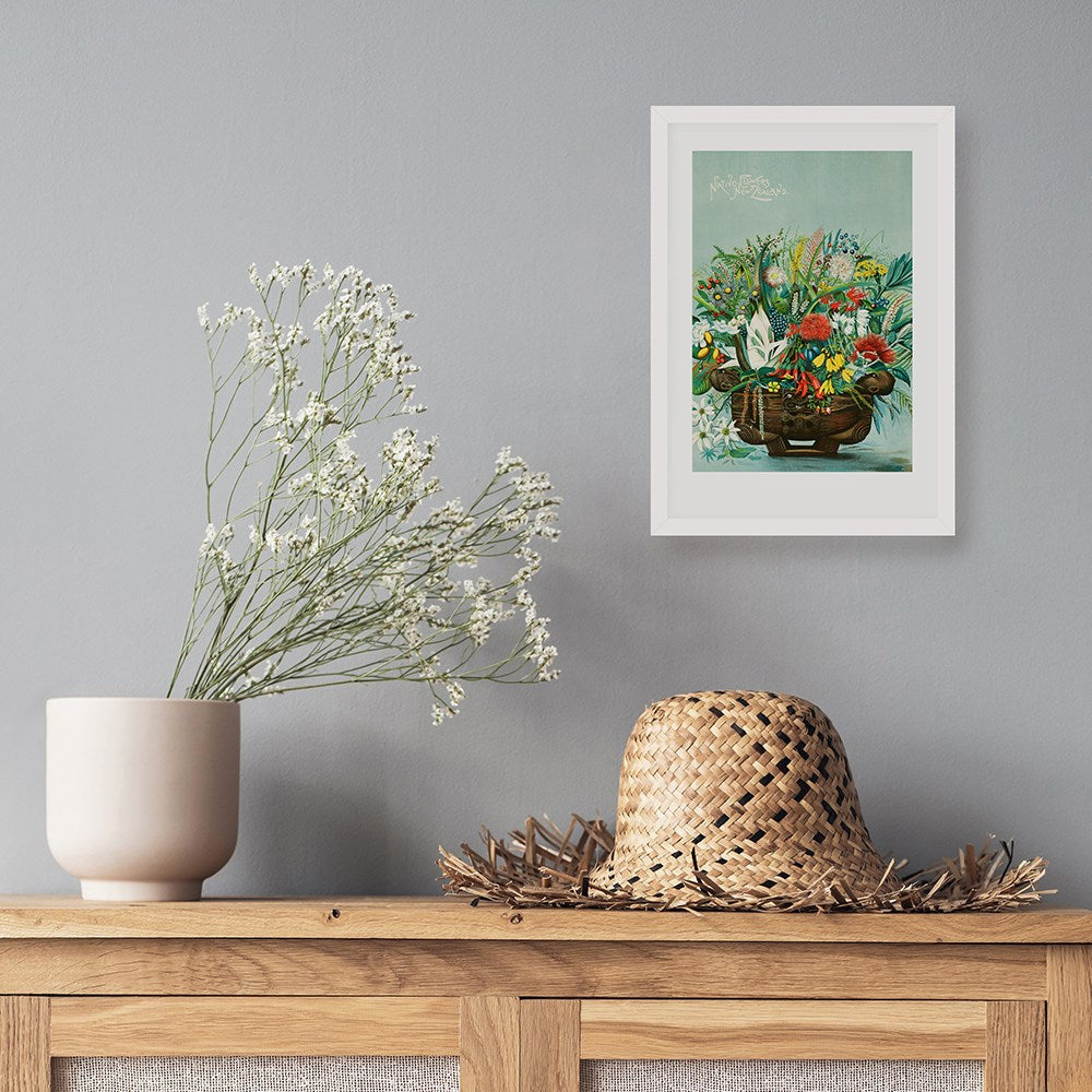 The NZ Native Flowers Print, a framed archival artwork of New Zealand flora, hangs on a gray wall above a wooden surface adorned with a white vase of flowers and a woven straw hat.