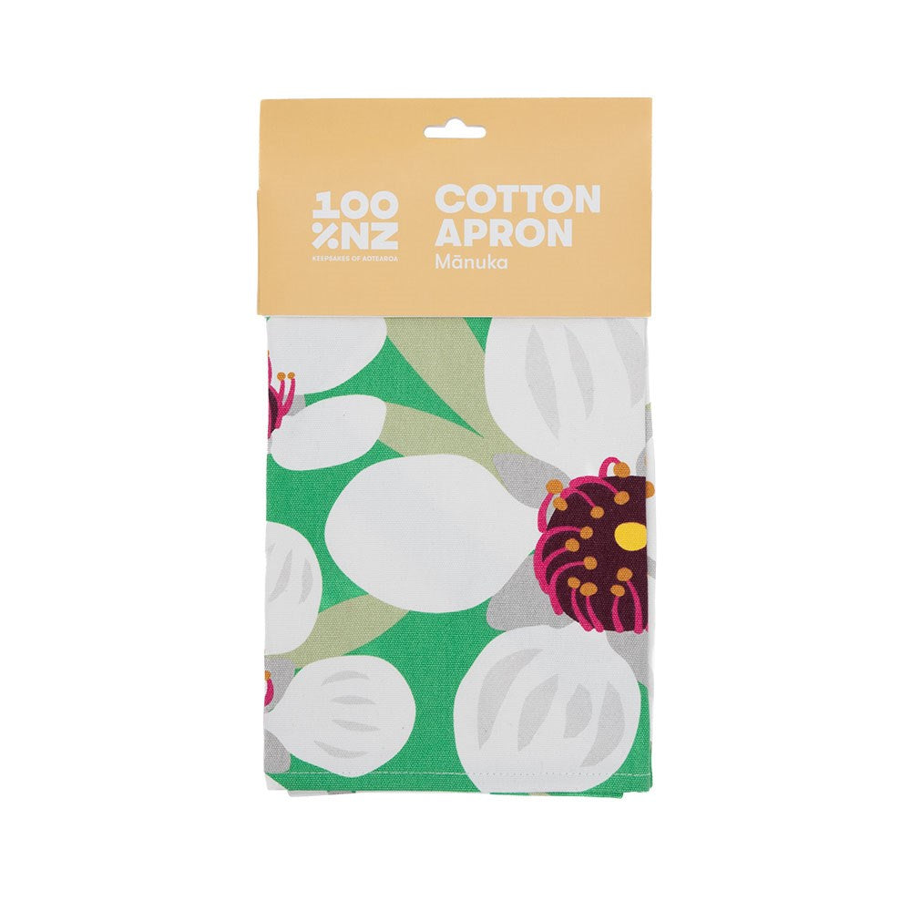 The Bright Botanical Manuka Apron is a folded 100% NZ cotton apron with a green, white, and purple floral design, packaged in a beige cardboard sleeve labeled 100% NZ Cotton Apron Mānuka and featuring distinctive New Zealand style.