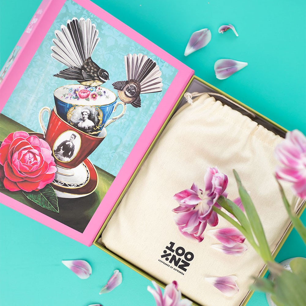 An open pink box featuring Angie Dennis’s “Deeds not Words” Artist Series jigsaw puzzle, with bird and teacup art on the lid, sits beside a cream "100% NZ" drawstring bag. Pink and white tulip petals rest on a turquoise surface.