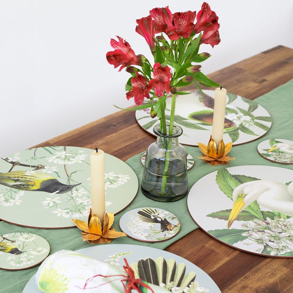 A wooden table set with a green runner, gold candle holders, red flowers in a glass vase, and Birds & Botanicals Placemats Set of 6—round cork-backed placemats and coasters featuring New Zealand birds and botanical illustrations.