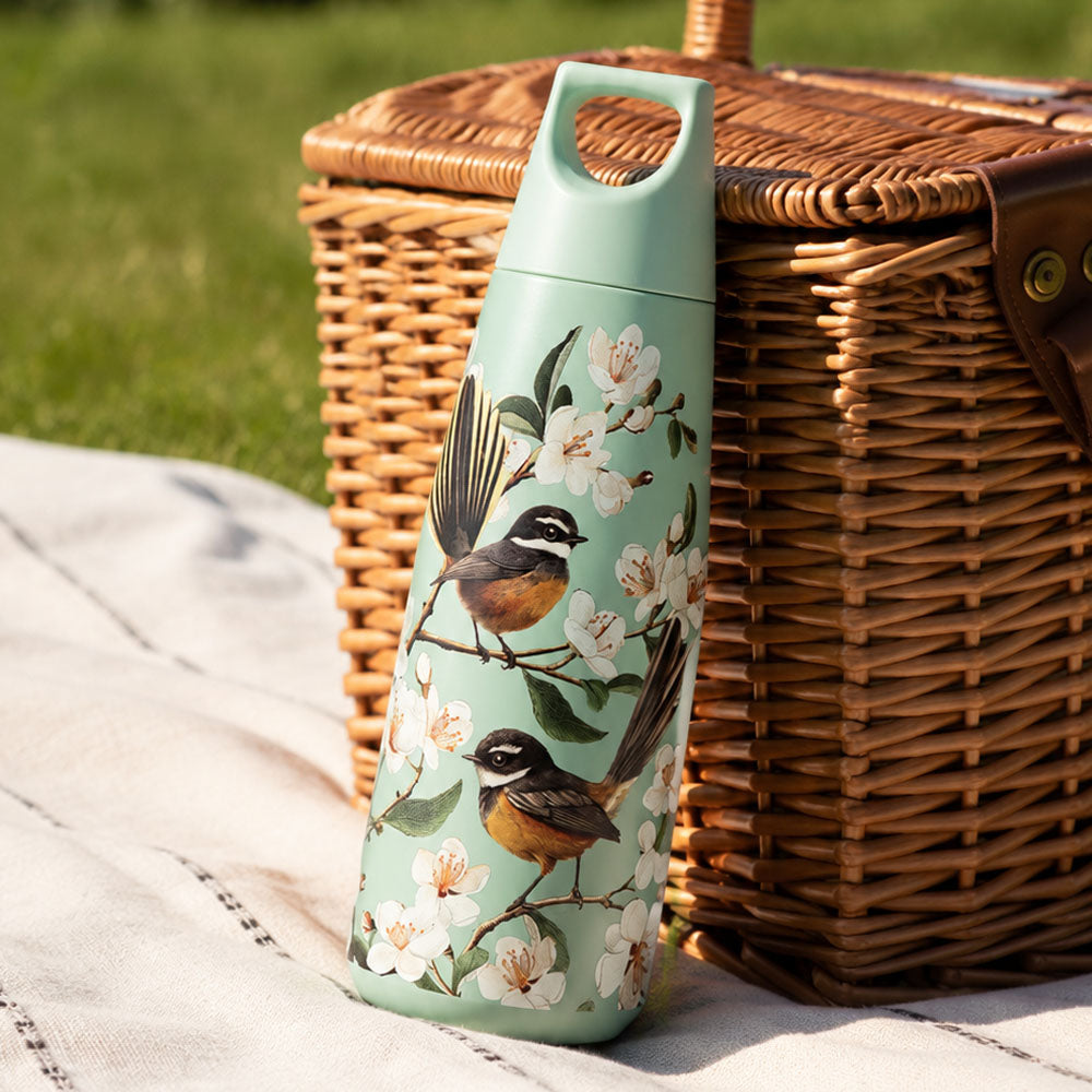 A hand holds the Pohutukawa Glade Drink Bottle—pink stainless steel with red floral patterns and a leak-resistant lid—near a pool, with lounge chairs and potted plants in the background.