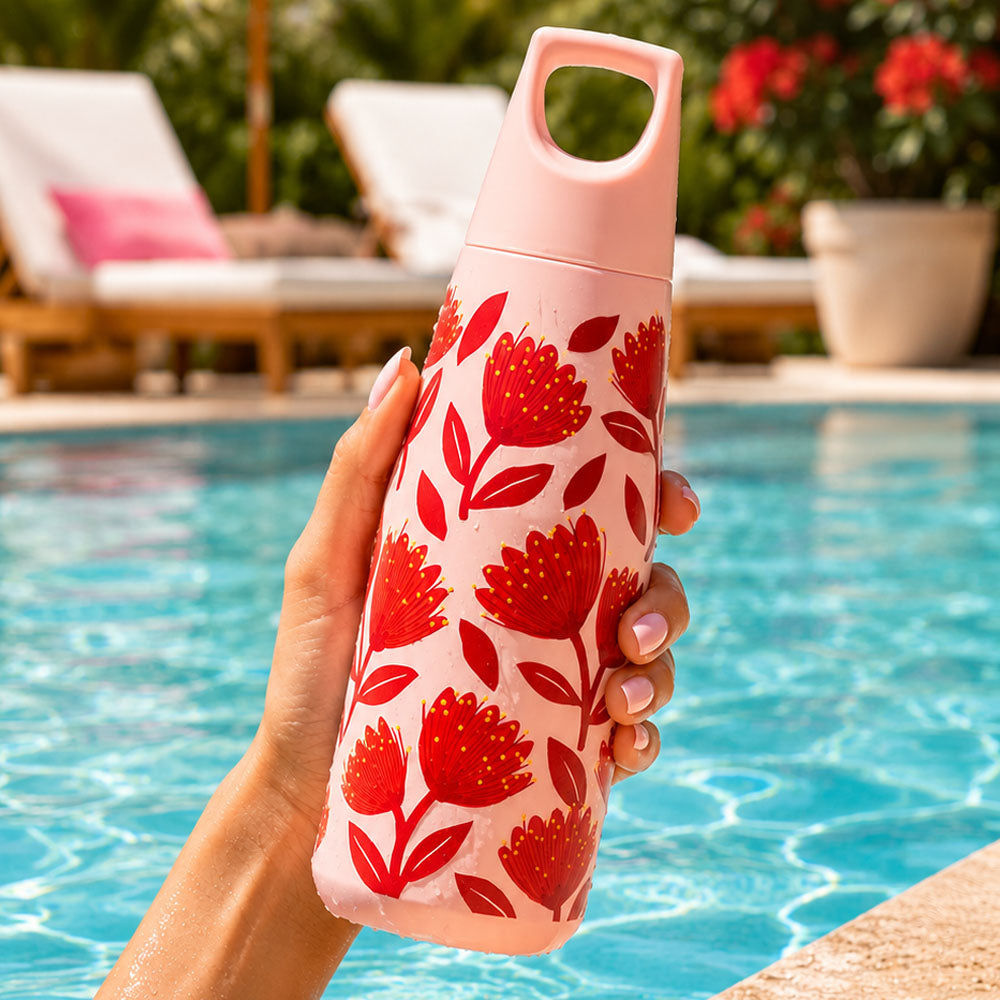 A hand holds the Pohutukawa Glade Drink Bottle—pink stainless steel with red floral patterns and a leak-resistant lid—near a pool, with lounge chairs and potted plants in the background.