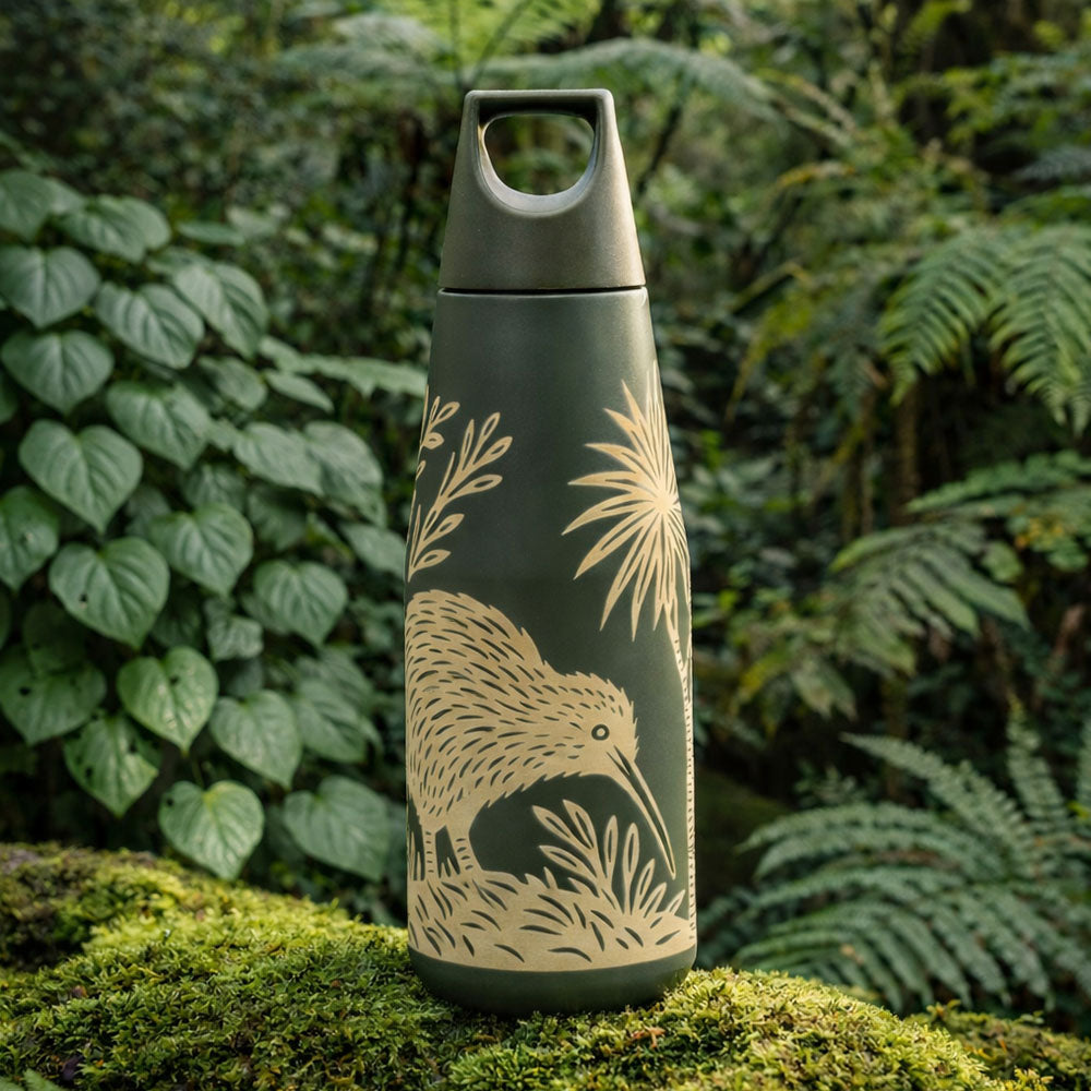 The Woodcut Kiwi Drink Bottle, a green vacuum insulated stainless steel bottle with a leak-resistant lid, stands on moss in a lush, leafy forest setting.