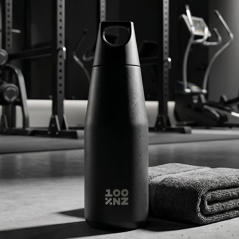The Obsidian Drink Bottle, a black BPA-free bottle featuring "100%NZ," stands upright beside a folded towel on a gym floor, with fitness equipment in the background.
