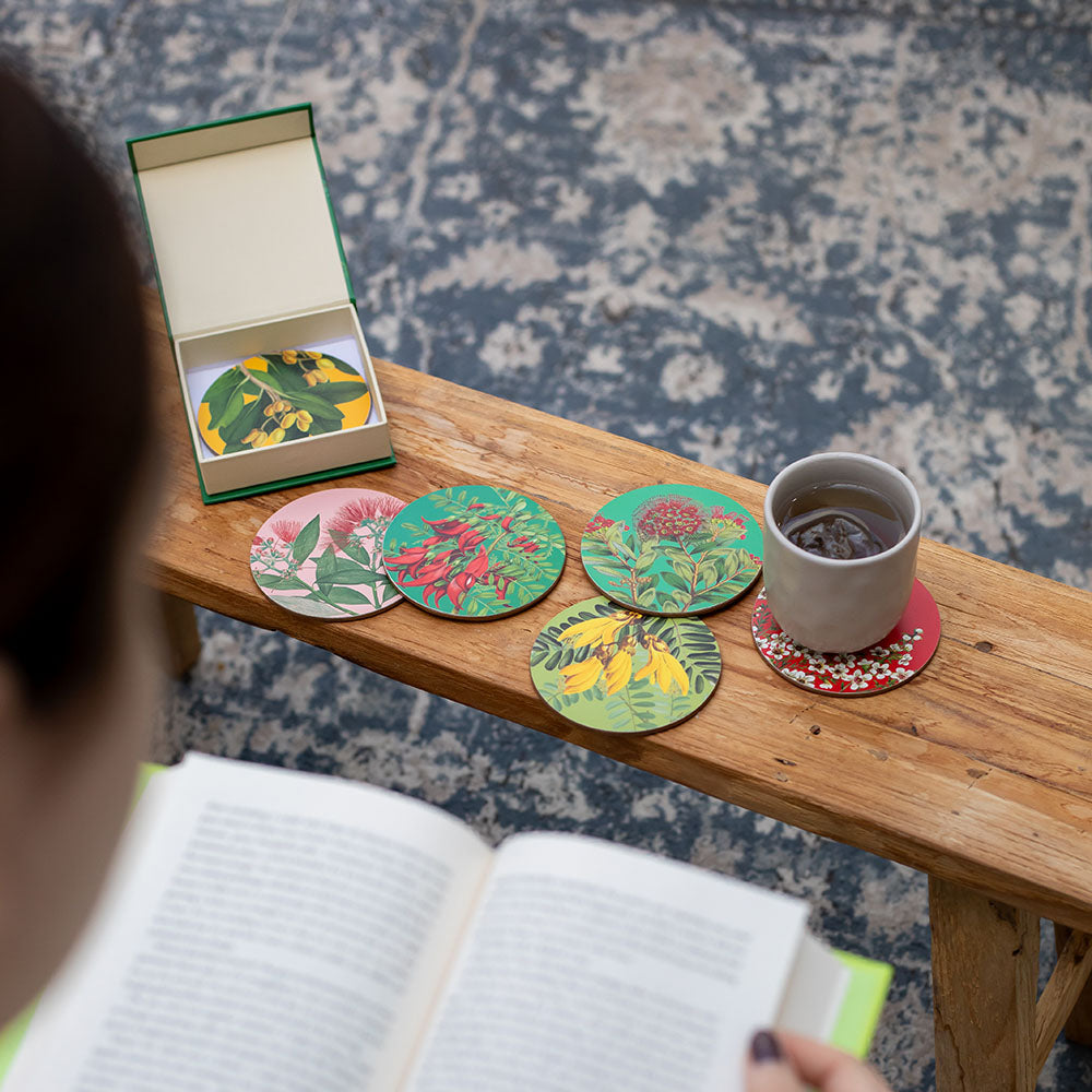 A person reads a book at a wooden table with coffee and several colorful Native Botanicals of NZ set of 6 Coasters, plus an open box of more coasters, all atop a patterned blue rug.