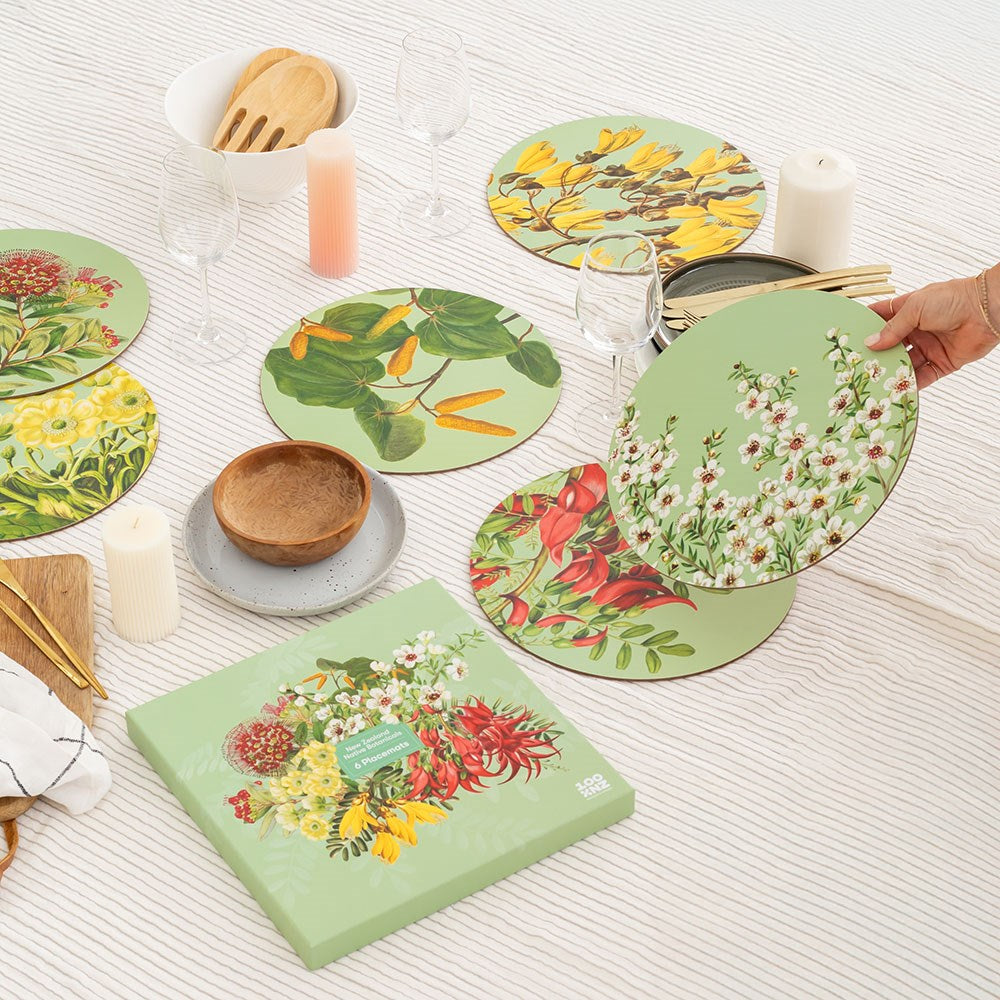 A hand arranges the NZ Native Botanicals Box of 6 Placemats, cork-backed and floral, on a white striped tablecloth. The table is set with glassware, candles, wooden utensils, stacked dishes, and a matching box.