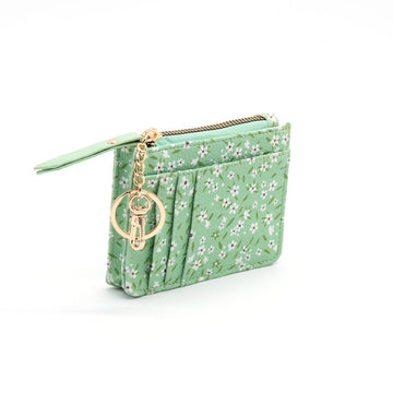 The Manuka Pattern Coin Purse is a small green vegan leather wallet with a white flower design, gold zipper, and keychain featuring a circular charm and key. It stands upright on a white background.