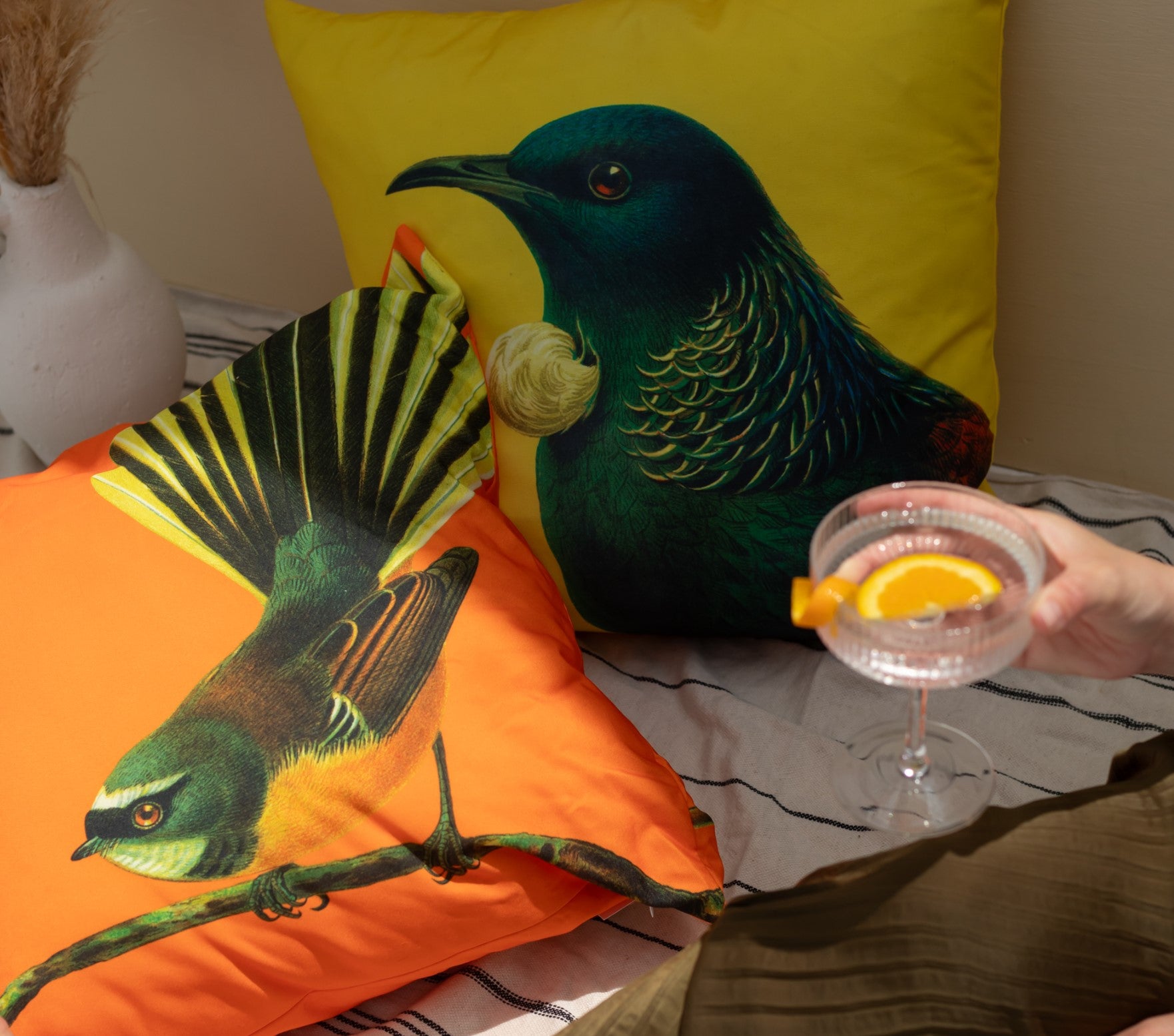 Two Bright Fantail Cushion Covers, adorned with colorful bird illustrations on polyester, rest on a striped surface. A hand holds a glass of iced orange drink; a white vase with dried grass sits in the background.