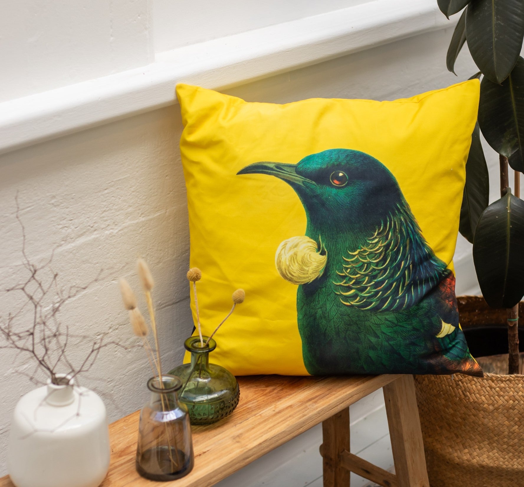 The Bright Tui Cushion Cover, a yellow polyester print designed in New Zealand, features a green and blue tui bird and is shown on a wooden bench with glass vases, dried plants, a basket, and an indoor plant.